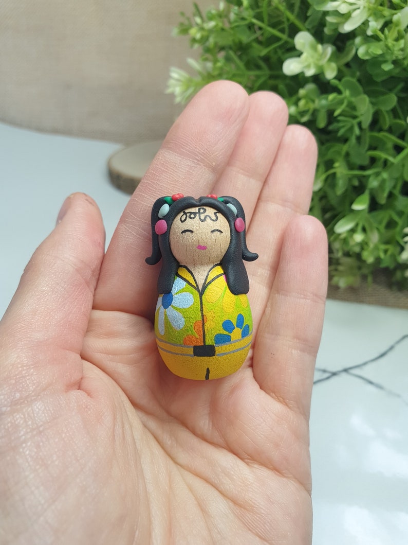 May include: A hand-painted wooden doll with black hair, a yellow shirt with a floral pattern, and a yellow skirt. The doll has a painted face with closed eyes and a small smile. The doll is wearing a black headband with red and white dots.