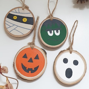 May include: Four wooden ornaments with Halloween themes. A mummy, a green monster, a pumpkin, and a ghost. Each ornament is painted with a different face and has a string for hanging.