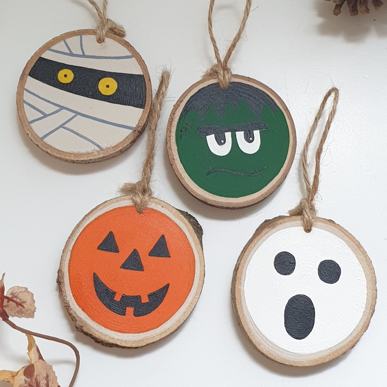 May include: Four wooden ornaments with Halloween themes. A mummy, a green monster, a pumpkin, and a ghost. Each ornament is painted with a different face and has a string for hanging.