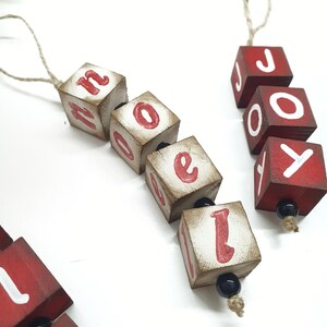 May include: Two wooden Christmas decorations spelling "NOEL" and "JOY" with red letters on a white background. The decorations are strung together with twine and black beads.