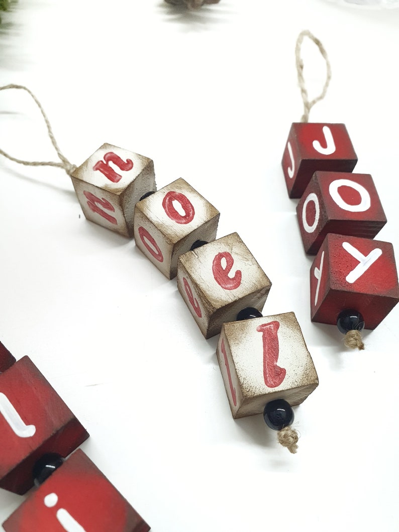May include: Two wooden Christmas decorations spelling "NOEL" and "JOY" with red letters on a white background. The decorations are strung together with twine and black beads.
