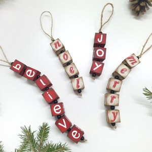 May include: Three red wooden Christmas decorations with white lettering spelling out "believe", "joy", and "merry". Each decoration is strung together with black beads and twine.