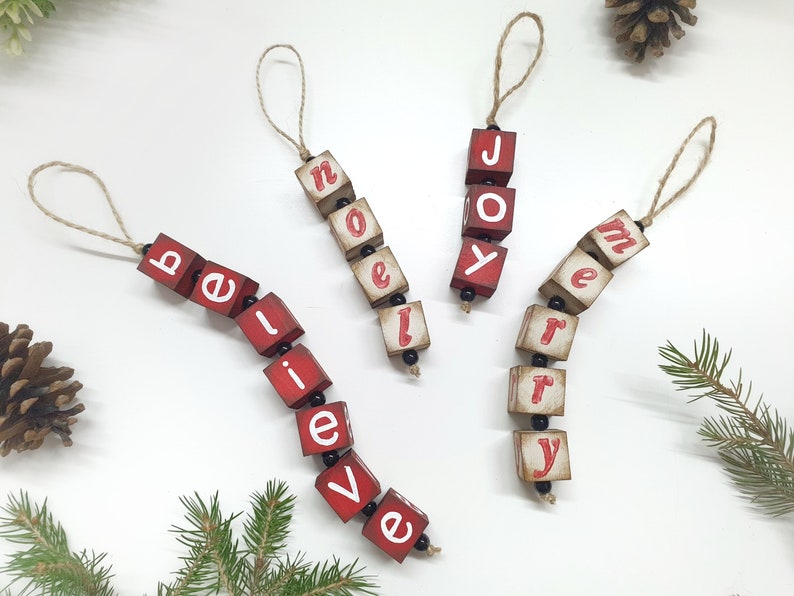 May include: Three red wooden Christmas decorations with white lettering spelling out "believe", "joy", and "merry". Each decoration is strung together with black beads and twine.