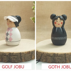May include: Two hand-painted wooden dolls, one dressed in a pink and gray golf outfit with a white visor and the other dressed in a black outfit with white accents and red hearts on the cheeks. The dolls are labeled "GOLF JOBU" and "GOTH JOBU".