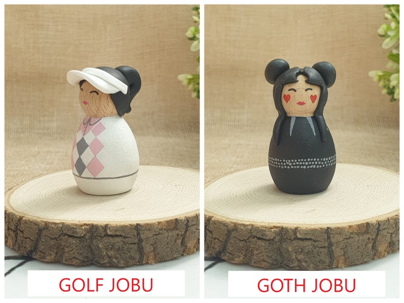 May include: Two hand-painted wooden dolls, one dressed in a pink and gray golf outfit with a white visor and the other dressed in a black outfit with white accents and red hearts on the cheeks. The dolls are labeled "GOLF JOBU" and "GOTH JOBU".