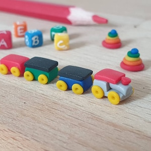 Puede incluir: Un tren de juguete con cuatro vagones, incluyendo una locomotora roja, un vagón verde, un vagón azul y un vagón gris. Los vagones tienen ruedas amarillas y están hechos de plástico. El tren está sobre una superficie de madera con bloques de letras de colores y anillos apilables.