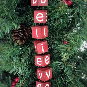 May include: Red wooden blocks spelling out the word "believe" hanging on a green artificial Christmas tree. The blocks are strung together with black beads.