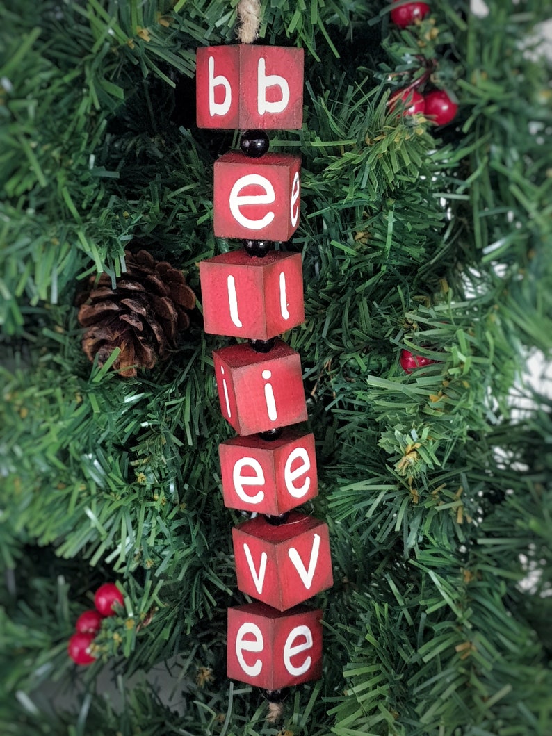 May include: Red wooden blocks spelling out the word "believe" hanging on a green artificial Christmas tree. The blocks are strung together with black beads.