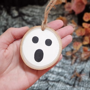 May include: A white wooden ghost ornament with black eyes and a mouth. The ornament is hanging from a twine string.