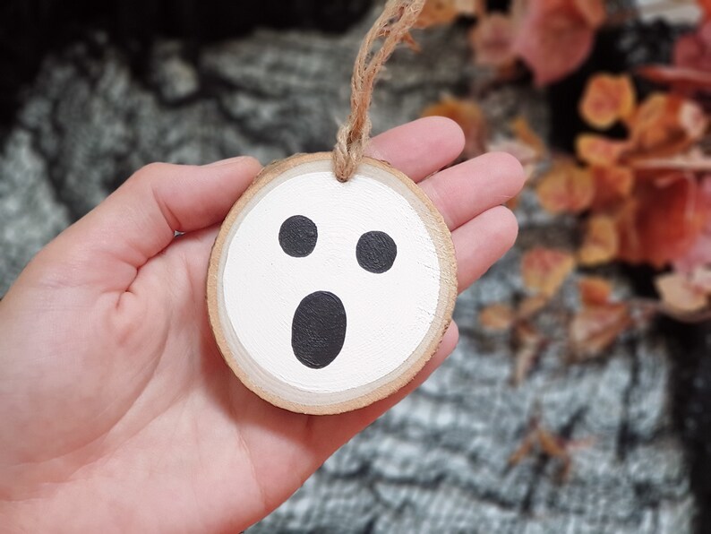 May include: A white wooden ghost ornament with black eyes and a mouth. The ornament is hanging from a twine string.