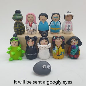 May include: A set of 11 hand-painted wooden figurines, each with unique features and designs. The figurines are arranged in two rows on a wooden surface. The figurines are all different colors and have different facial expressions. There is also a small gray rock with googly eyes in the bottom center of the image. It will be sent a googly eyes rock pin to those who buy the 5 or 10 Jobu set!