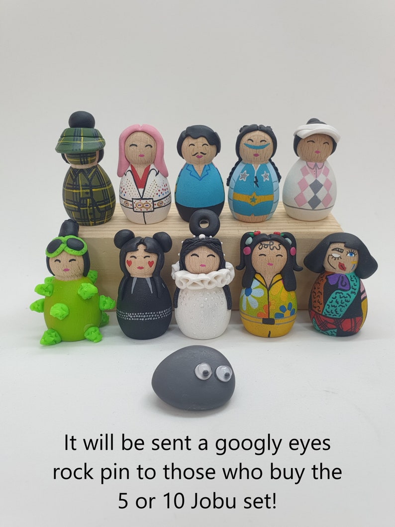 May include: A set of 11 hand-painted wooden figurines, each with unique features and designs. The figurines are arranged in two rows on a wooden surface. The figurines are all different colors and have different facial expressions. There is also a small gray rock with googly eyes in the bottom center of the image. It will be sent a googly eyes rock pin to those who buy the 5 or 10 Jobu set!