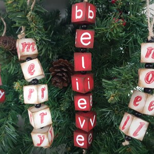 May include: Four wooden Christmas decorations spelling out "Merry", "Believe", "Noel", and "Joy" with red letters and black beads.