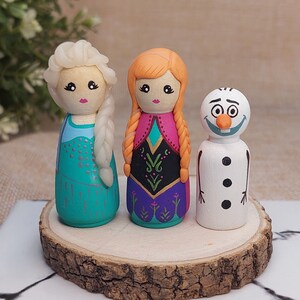 May include: Three hand-painted wooden peg dolls depicting characters from the film Frozen. The dolls are Elsa with a light blue dress and white hair, Anna with a pink and purple dress and red hair, and Olaf the snowman with a white body and orange nose.