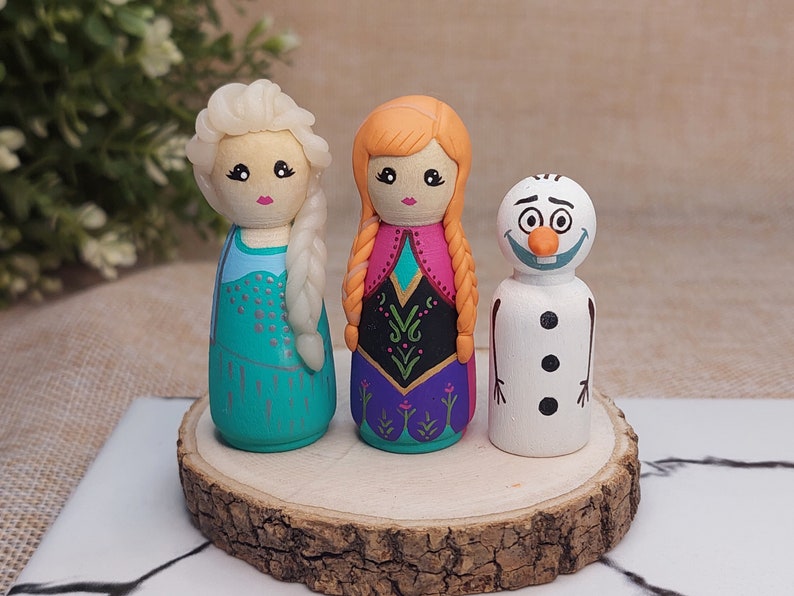 May include: Three hand-painted wooden peg dolls depicting characters from the film Frozen. The dolls are Elsa with a light blue dress and white hair, Anna with a pink and purple dress and red hair, and Olaf the snowman with a white body and orange nose.