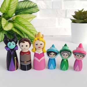 Puede incluir: Un conjunto de siete muñecos de madera pintados a mano con personajes de La Bella Durmiente de Disney. Los muñecos están vestidos con sus trajes icónicos, incluyendo Maléfica con un vestido morado, el Príncipe Felipe con un atuendo azul y dorado, Aurora con un vestido rosa y las tres hadas con vestidos verdes, azules y rosas.