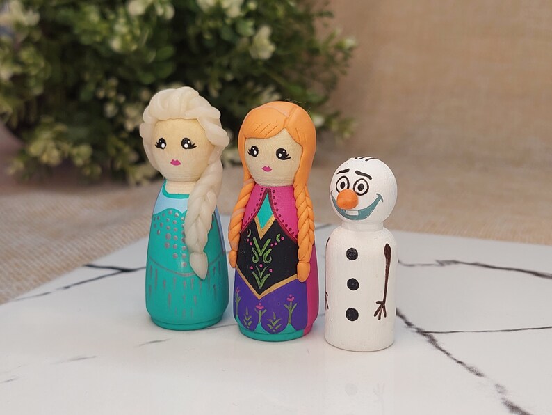 May include: Three wooden peg dolls, painted to resemble characters from the film Frozen. The dolls are Elsa, Anna, and Olaf. Elsa is wearing a blue and silver dress with a blonde braid. Anna is wearing a purple and pink dress with a red braid. Olaf is wearing a white and orange outfit with a black smile.