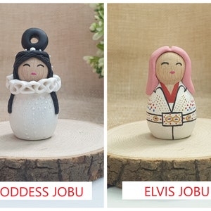 May include: Two handmade wooden dolls, one with a white dress and black hair, the other with a pink wig and a white jumpsuit with gold accents. The dolls are labeled "Goddess Jobu" and "Elvis Jobu".