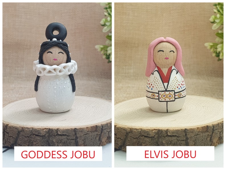May include: Two handmade wooden dolls, one with a white dress and black hair, the other with a pink wig and a white jumpsuit with gold accents. The dolls are labeled "Goddess Jobu" and "Elvis Jobu".