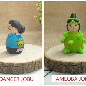 May include: Two hand-painted wooden figurines. The first figurine is a dancer with a blue dress and a colorful scarf. The second figurine is an amoeba with green and black details. Both figurines are on wooden bases.