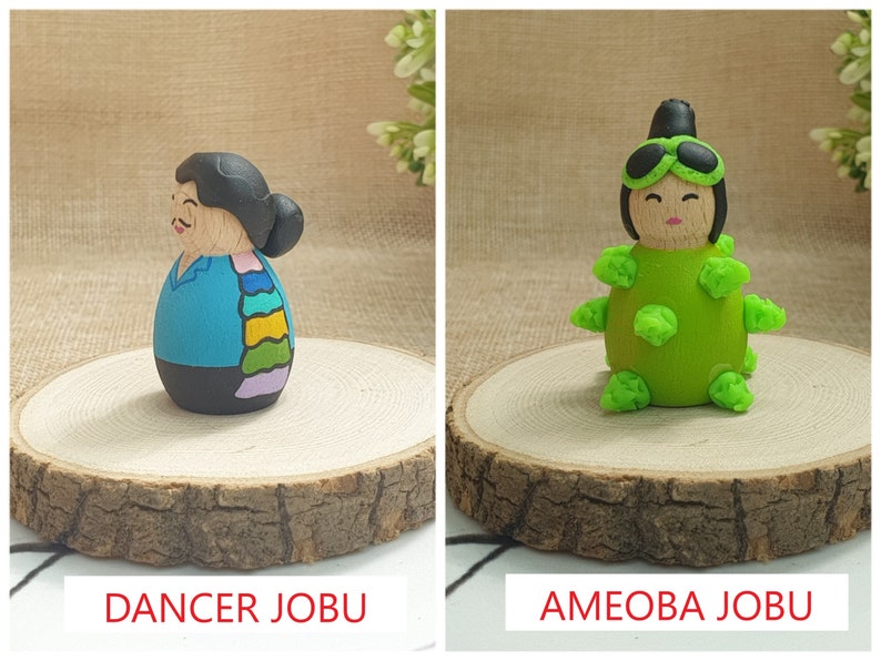 May include: Two hand-painted wooden figurines. The first figurine is a dancer with a blue dress and a colorful scarf. The second figurine is an amoeba with green and black details. Both figurines are on wooden bases.