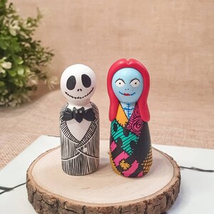Könnte beinhalten: Zwei handbemalte Holzfiguren von Jack Skellington und Sally aus dem Film Nightmare Before Christmas. Die Figuren stehen auf einer Holzscheibe.