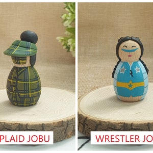 May include: Two wooden doll figures, one with a green plaid hat and jacket, the other with a blue and yellow wrestling costume. The figures are standing on wooden bases. The text "PLAID JOBU" is below the first figure and "WRESTLER JOBU" is below the second figure.