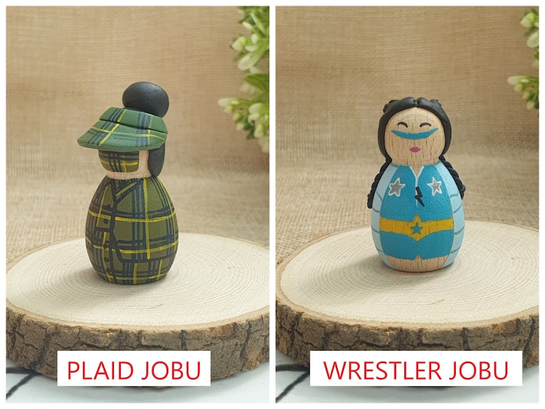 May include: Two wooden doll figures, one with a green plaid hat and jacket, the other with a blue and yellow wrestling costume. The figures are standing on wooden bases. The text "PLAID JOBU" is below the first figure and "WRESTLER JOBU" is below the second figure.