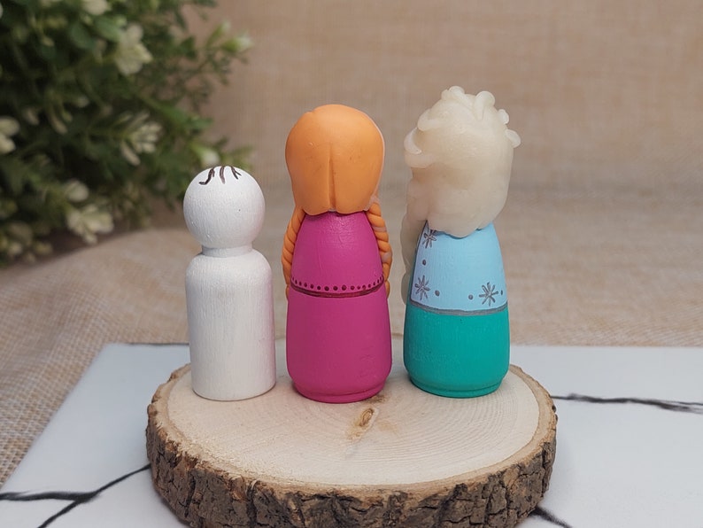 May include: Three wooden peg dolls, painted to resemble characters from the film Frozen. The doll on the left is white, the middle doll is pink with orange hair, and the doll on the right is blue and green with white hair.
