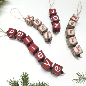 May include: Three red wooden ornaments with white letters spelling out "believe", "joy", and "merry". Each ornament is strung together with twine.