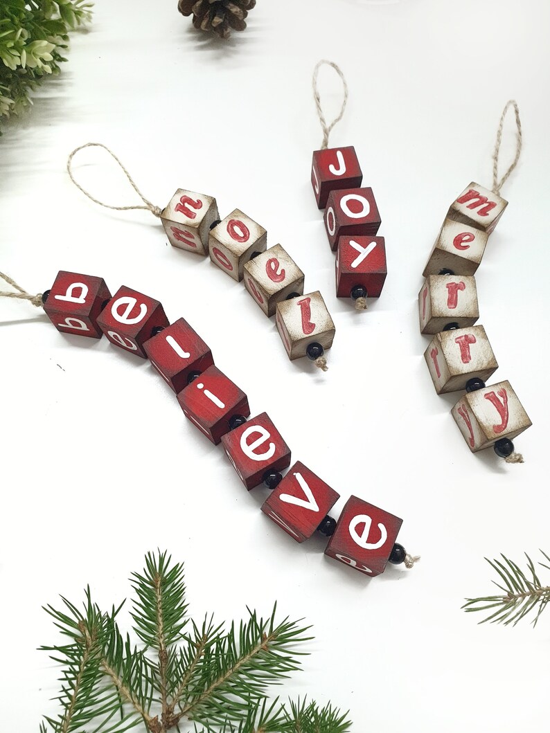 May include: Three red wooden ornaments with white letters spelling out "believe", "joy", and "merry". Each ornament is strung together with twine.