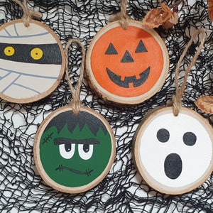 May include: Four wooden Halloween ornaments with painted faces. A mummy with yellow eyes, a pumpkin with a jack-o-lantern face, a green Frankenstein monster with stitches, and a white ghost with black eyes and a mouth.