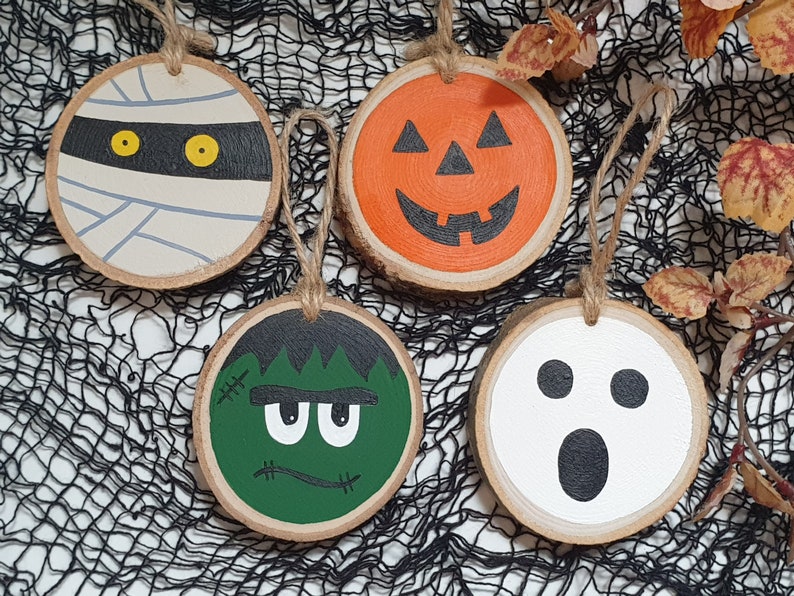 May include: Four wooden Halloween ornaments with painted faces. A mummy with yellow eyes, a pumpkin with a jack-o-lantern face, a green Frankenstein monster with stitches, and a white ghost with black eyes and a mouth.