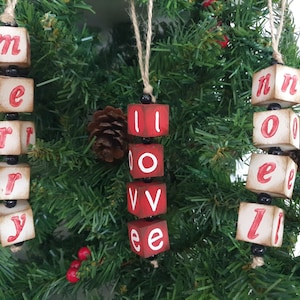 May include: Three wooden Christmas decorations spelling out "Merry", "Love", and "Noel". Each decoration is made of individual blocks with red letters on a white background. The blocks are strung together with black beads.