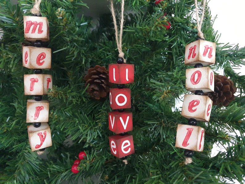May include: Three wooden Christmas decorations spelling out "Merry", "Love", and "Noel". Each decoration is made of individual blocks with red letters on a white background. The blocks are strung together with black beads.