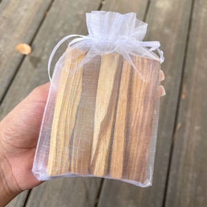 May include: A bundle of light brown Palo Santo sticks, held in a sheer white organza bag with a drawstring closure. The wood has a natural grain and varying shades of brown and tan. The bag is tied at the top.