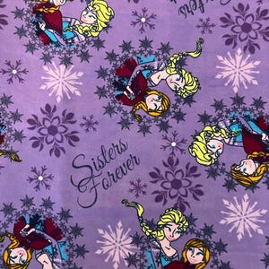 May include: Purple fabric with a repeating pattern of two sisters, Anna and Elsa from the movie Frozen, hugging each other. The fabric has a snowflake and floral pattern and the text "Sisters Forever".