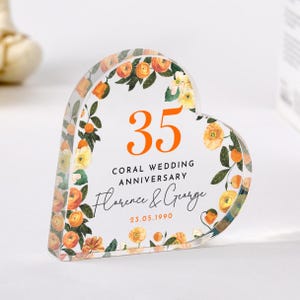 Könnte beinhalten: Ein herzförmiger, transparenter Acryl-Briefbeschwerer mit einem floralen Design in Orange- und Gelbtönen. Die Zahl 35 ist in Orange in der Mitte des Briefbeschwerers gedruckt. Der Text "Coral Wedding Anniversary Florence & George 23.05.1990" ist unter der Zahl gedruckt.