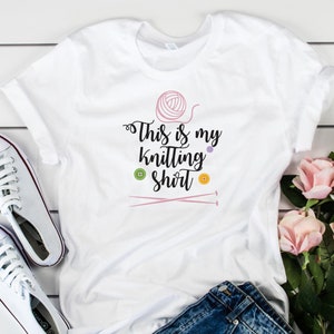 Pu&ograve; includere: Maglietta bianca con il testo "This is my knitting shirt" in lettere nere. La maglietta presenta un gomitolo di lana rosa, due ferri da maglia rosa e tre bottoni colorati.