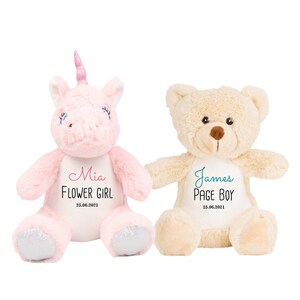 May include: Two plush toys, a pink unicorn and a beige teddy bear, both wearing white shirts with personalized text. The unicorn's shirt says "Mia Flower Girl 25.06.2021" in pink and black lettering. The teddy bear's shirt says "James Page Boy 25.06.2021" in teal and black lettering.
