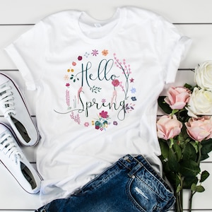 Puede incluir: Camiseta blanca con un diseño floral y el texto "Hello Spring" en un círculo.