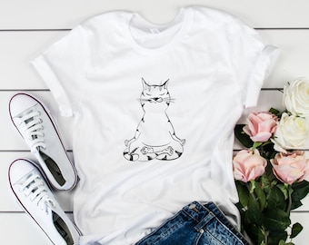 May include: White t-shirt with a black line drawing of a cat sitting in a yoga pose. The cat has its eyes closed and is meditating. The t-shirt is laid out on a white surface with a pair of white sneakers and blue denim shorts.