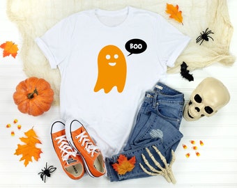 May include: White t-shirt featuring an orange ghost graphic and the word "BOO" in a black speech bubble. The shirt is displayed on a white surface with autumnal decorations, including a pumpkin, leaves, and candy corn.