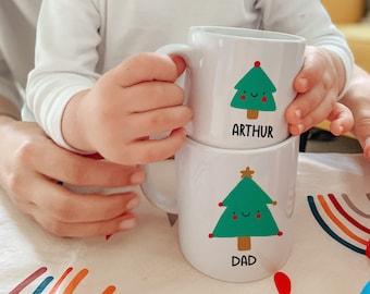 Könnte beinhalten: Zwei weiße Keramikbecher mit Weihnachtsbaummotiven. Der obere Becher hat einen grünen Baum mit einem roten Stern und dem Text "ARTHUR". Der untere Becher hat einen grünen Baum mit einem goldenen Stern und dem Text "DAD".