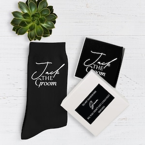 May include: Black socks with the text "Jack the Groom" in white script. A gift box with the same text and a label that reads "To my handsome, so you don't get cold feet 01.05.2022". A succulent plant is also in the image.