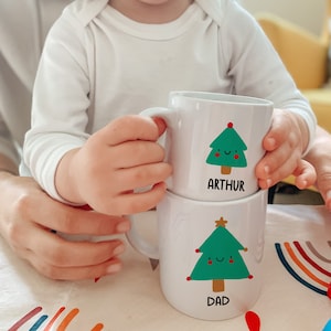 May include: Two white ceramic mugs with Christmas tree designs. The top mug has a green tree with a red star and the text "ARTHUR". The bottom mug has a green tree with a gold star and the text "DAD".