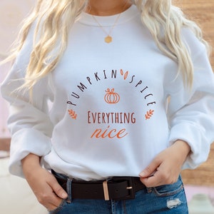 Peut inclure: Sweat-shirt blanc avec le texte "Pumpkin Spice Everything Nice" en lettres noires et orange. Un petit graphique de citrouille orange est centré au-dessus du texte.
