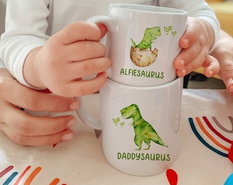 May include: Two white ceramic mugs with green dinosaur illustrations. The top mug says "Alfiesaurus" and the bottom mug says "Daddysaurus".
