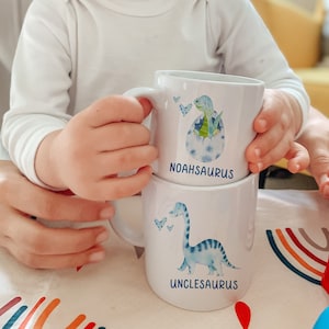 Pu&ograve; includere: Due tazze di ceramica bianca impilate. La tazza superiore presenta un dinosauro blu che esce da un uovo, con il nome "NOAHSAURUS" sotto. La tazza inferiore ha un dinosauro blu con il nome "UNCLESAURUS".