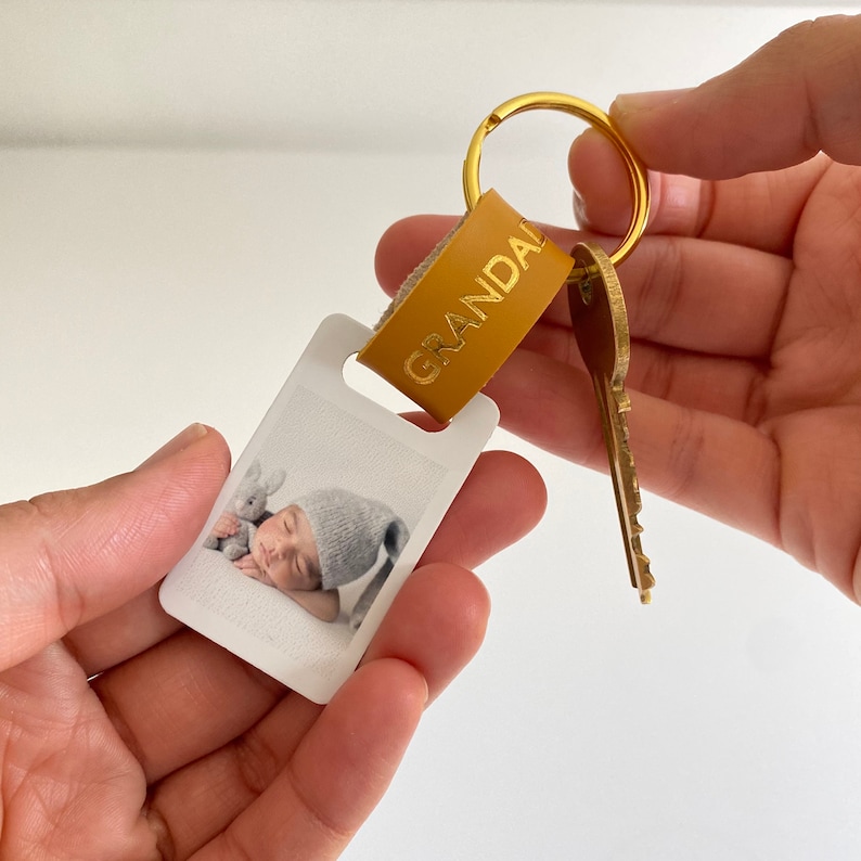 Puede incluir: Un llavero de foto blanco con una imagen de un beb&eacute; durmiendo. El llavero tiene una correa de cuero marr&oacute;n con la palabra "GRANDAL" grabada en letras doradas.